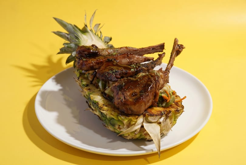 Grilled lamb chops with sauce and herbs, served in a pineapple half on a white plate.