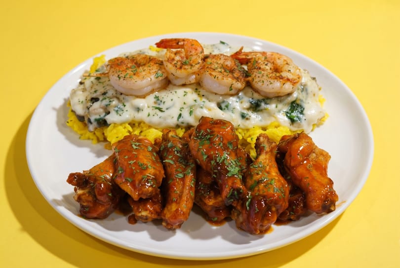 A white plate with yellow rice, creamy spinach, seasoned shrimp, and BBQ chicken wings.